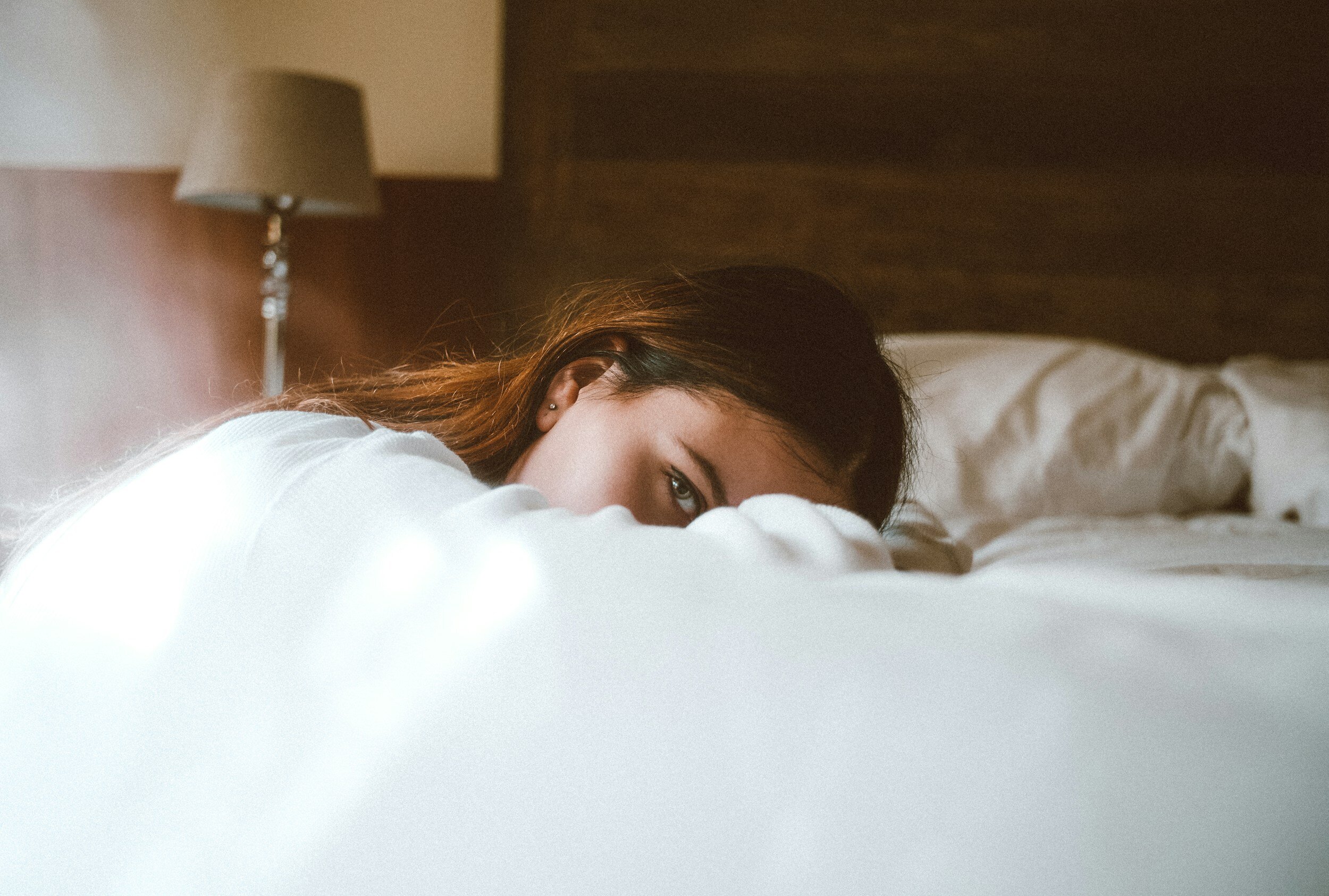 long-haired woman lying in a bed, looking in the camera, half of her face covered with linen.