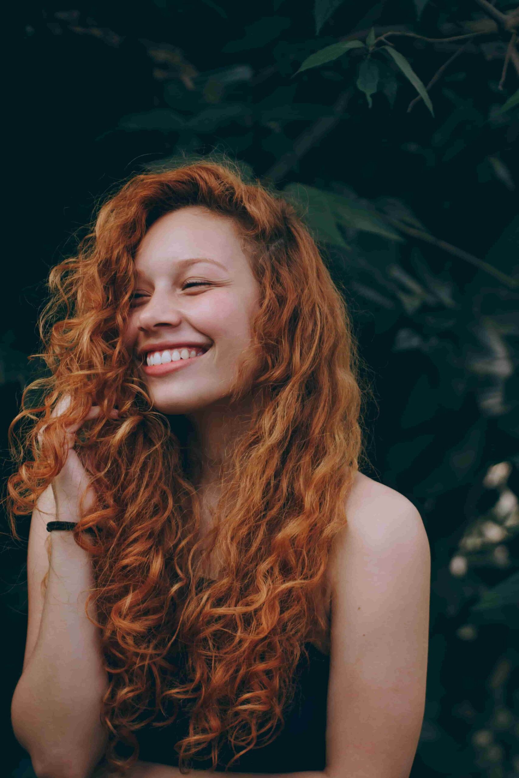 Woman with gorgeous, curly, red hair smiling in front of green plants