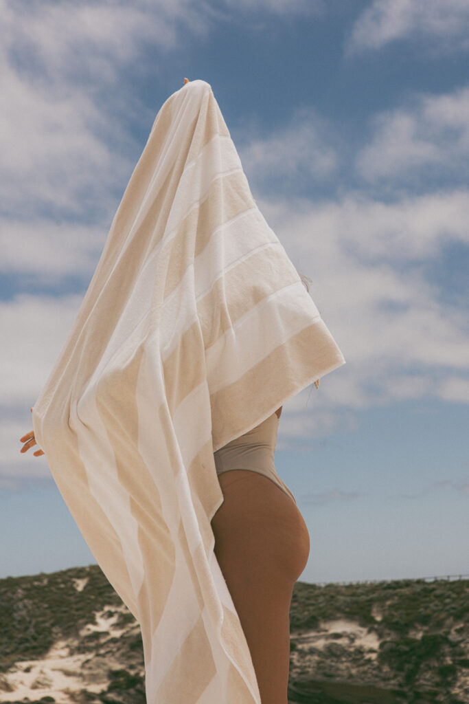 Woman on a beach, standing up, her face behind a striped beach towel. 