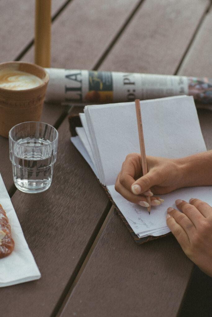 Close-up to a hand writing on a white notebook. Next to it, on the table, a latte, a glass of water, and a rolled newspaper. 