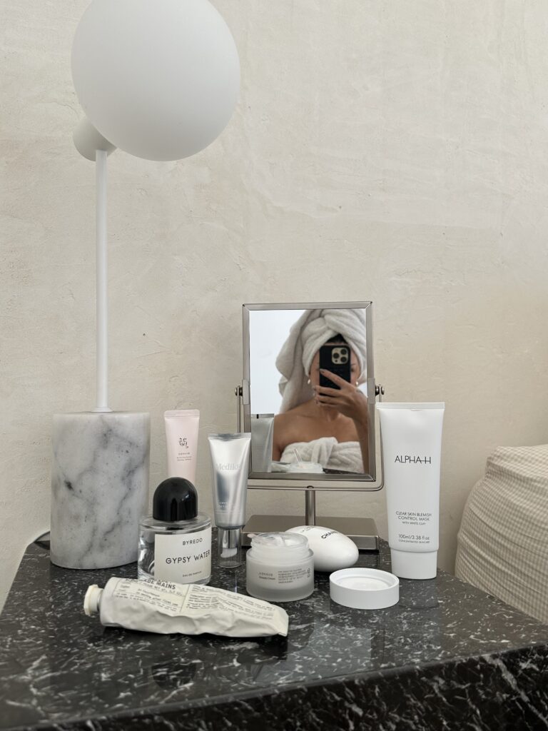 A serene morning scene shows a neatly arranged vanity table in a softly lit bedroom. A small mirror reflects the surroundings while various skincare products are artfully placed. A marble container holds beauty essentials, and a lamp adds gentle illumination. A towel-wrapped figure is partially visible in the mirror, suggesting a fresh start to the day. Might be following the Ayurvedic morning routine of easing into the day. 