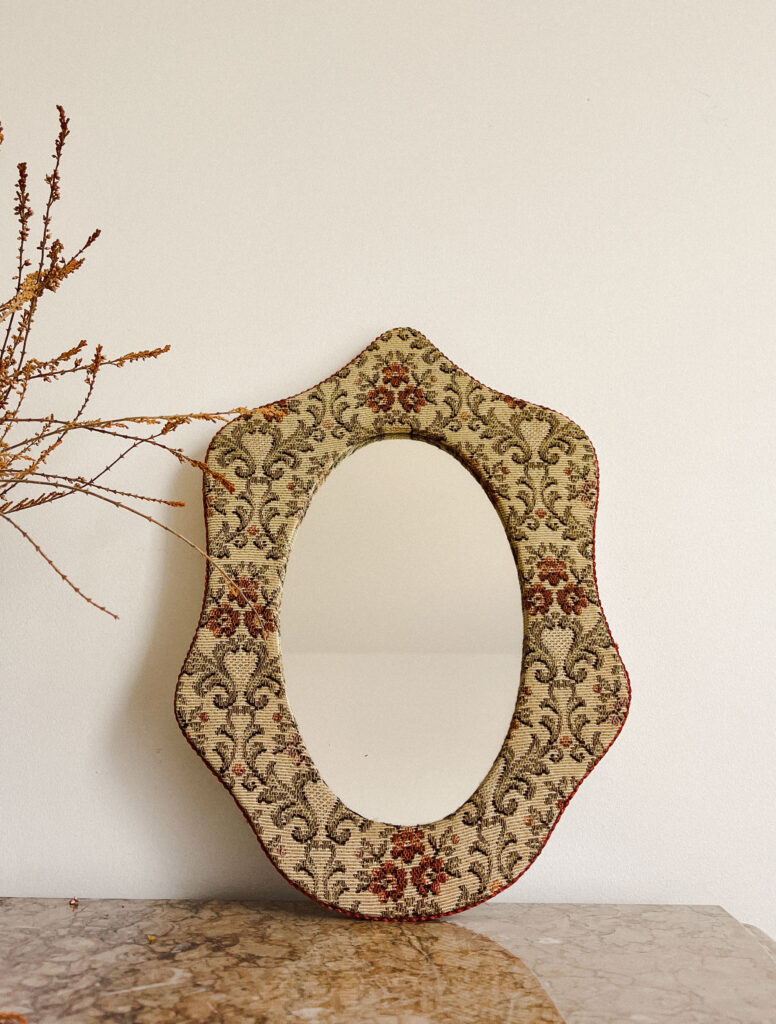Image of an unusal-shaped mirror, with oriental-looking motifs, next to what seems to be fake flowers. 