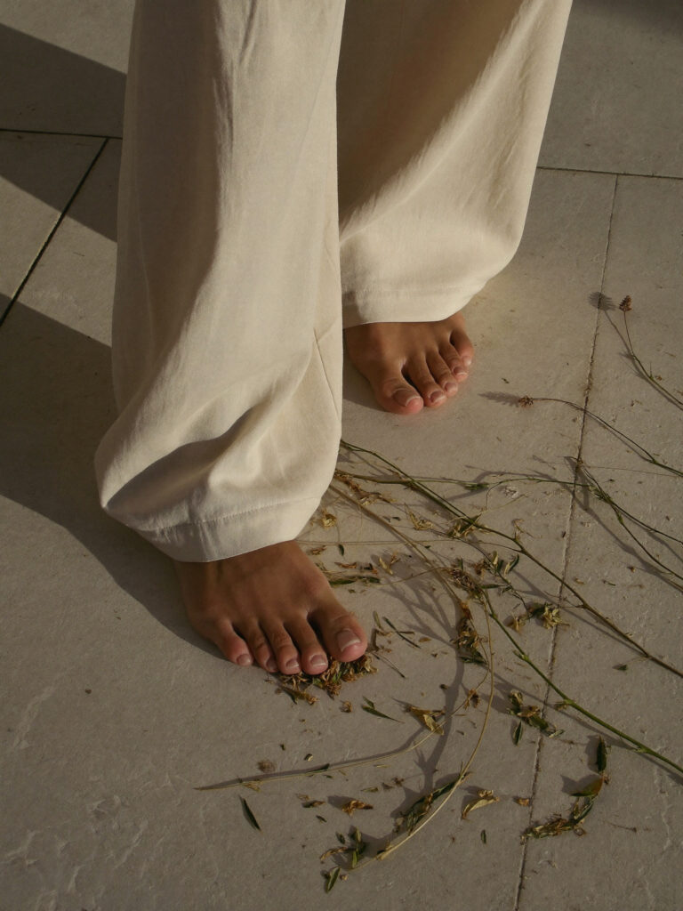 Barefoot woman in cream wide-leg trousers standing on pale stone tiles among scattered wild flowers