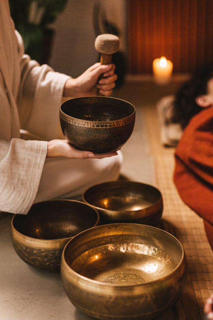 Person in cream linen holding a Tibetan singing bowl and mallet while seated. Sound healing is an ally of your nervous system